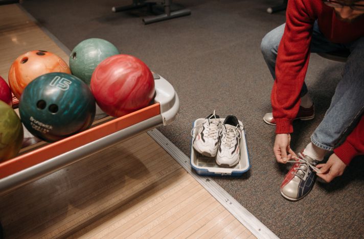 Eine Person schnürt Bowlingschuhe neben einer Ablage mit Bowlingkugeln